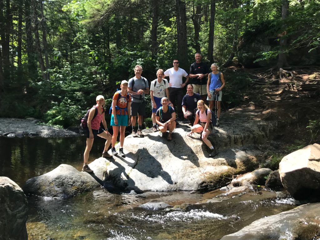 10 people gather on a large rock in the stream for a group photo during a Guild Gathering in the NE U.S. in 2019